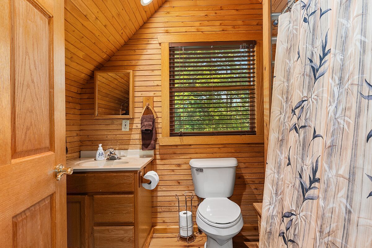Une salle de bain rustique, aménagée dans une cabane en bois, avec des toilettes, un lavabo et une fenêtre équipée de stores.