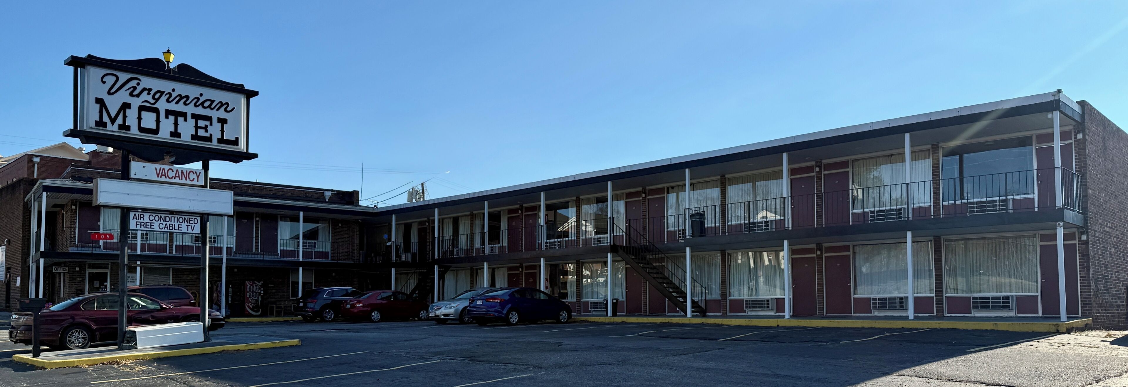 Un motel con un letrero que dice 'Virginian MOTEL' y un letrero de 'VACANCY'. Hay coches estacionados al frente, y el edificio tiene varios pisos con ventanas y balcones.