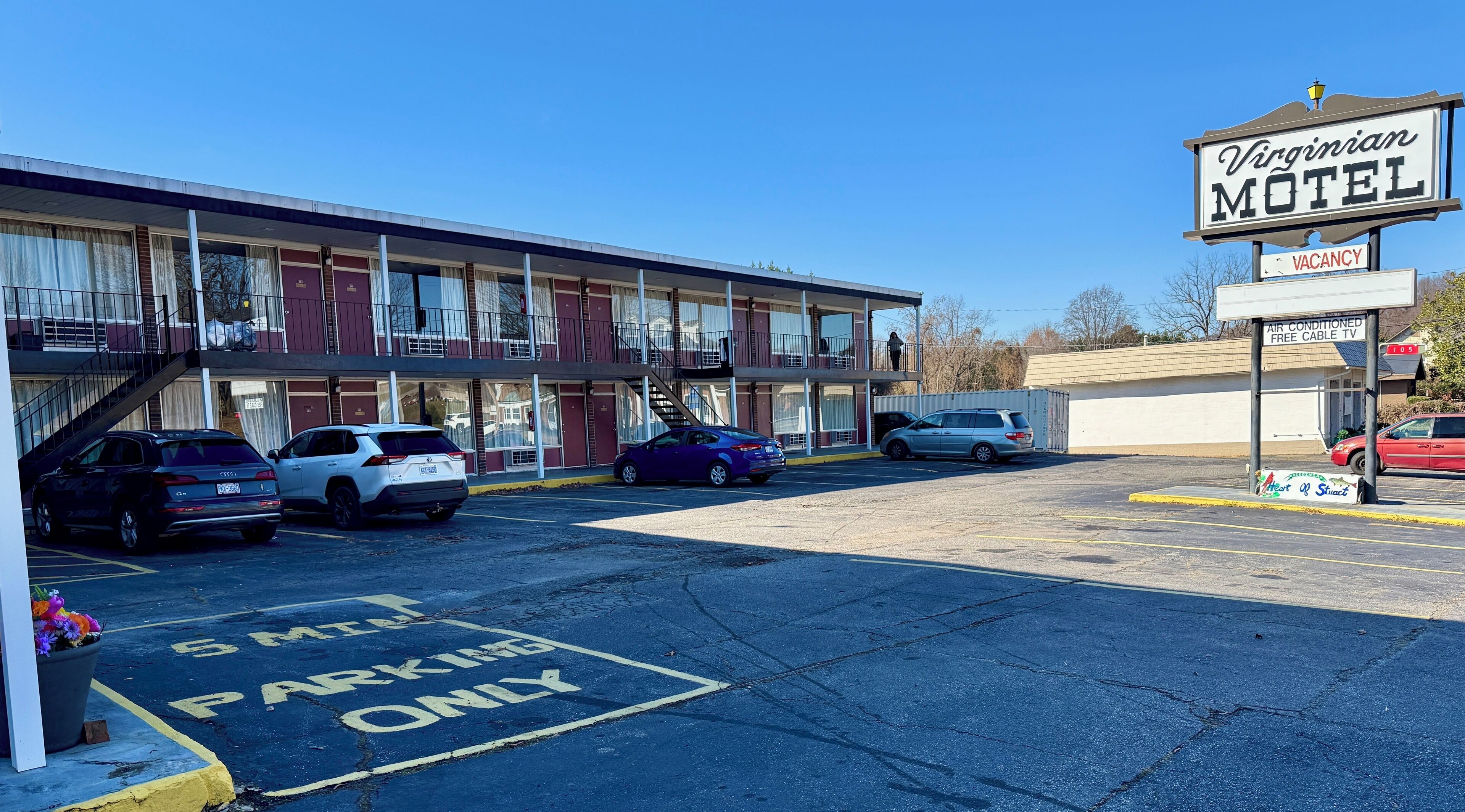 Un motel con un cartel que indica habitaciones disponibles, un estacionamiento con espacios delimitados y un edificio con múltiples ventanas y balcones.