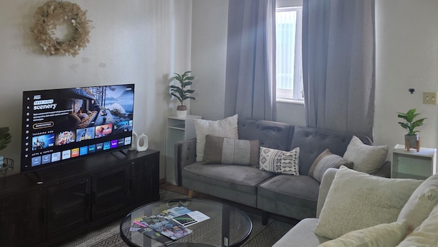 Un salon équipé d’un téléviseur à écran plat, d’un ventilateur de plafond, d’un canapé gris, d’une table basse et d’un tapis à motifs.