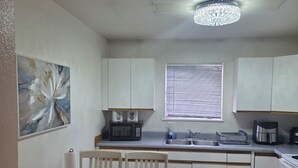 Une cuisine avec des meubles blancs, une table à manger avec des chaises, un four à micro-ondes, une machine à café et une fenêtre avec des stores.