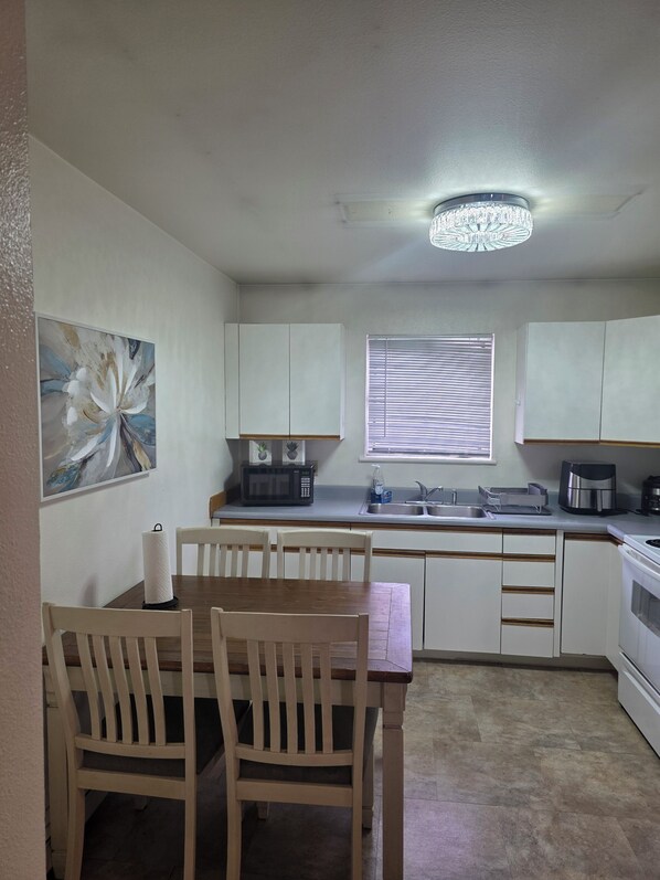A kitchen with white cabinets, a dining table with chairs, a microwave, a coffee maker, and a window with blinds.