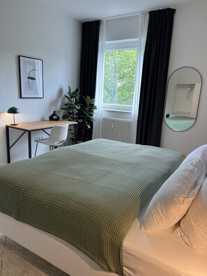 Ein ordentlich eingerichtetes Schlafzimmer mit Bett, Schreibtisch, Stuhl, Topfpflanze, einem Fenster mit Vorhängen und einem Spiegel an der Wand.