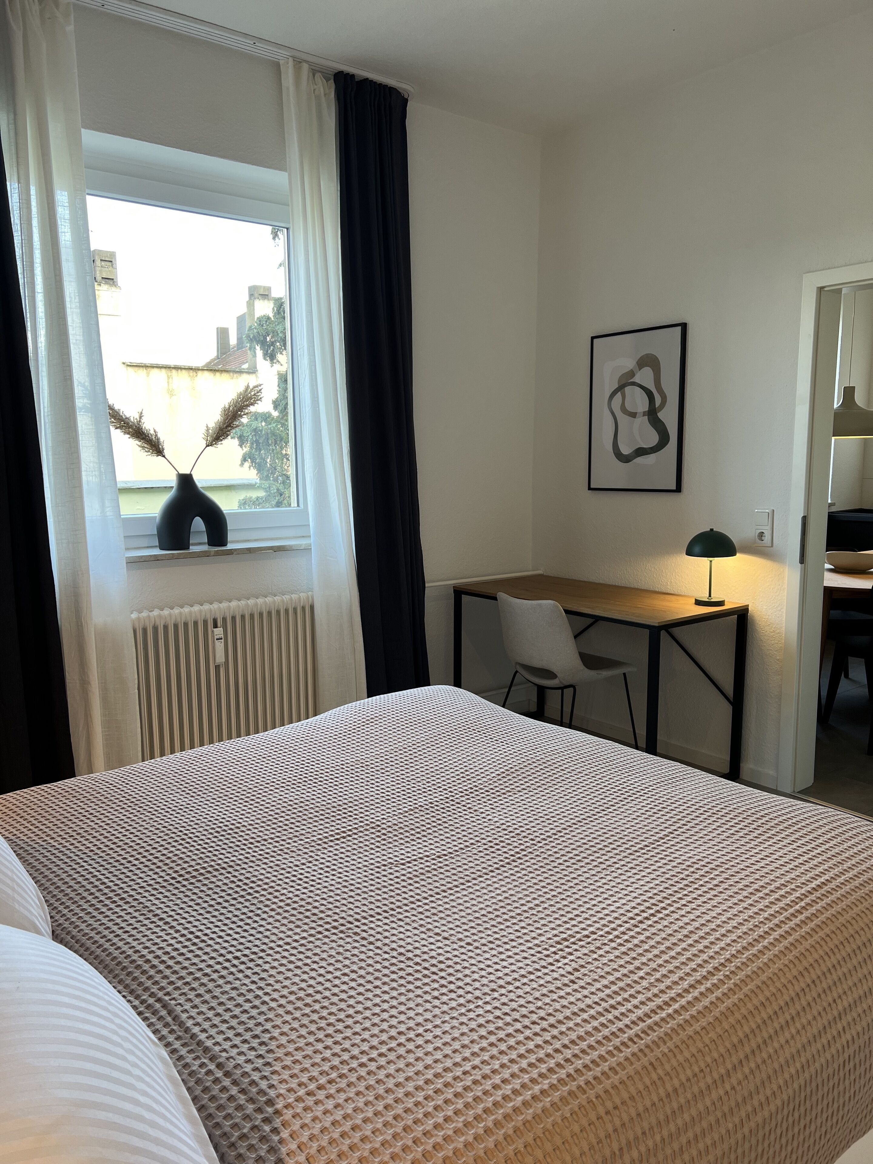 Habitación de hotel con cama, escritorio, silla, radiador y ventana con cortinas.