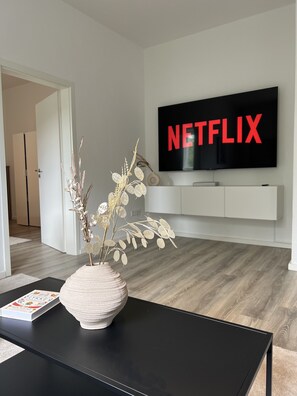 Un salon moderne avec une table basse noire, un vase blanc contenant des plantes séchées, un téléviseur à écran plat fixé au mur qui affiche le logo « NETFLIX », et un meuble blanc pour équipements audiovisuels placé en dessous.