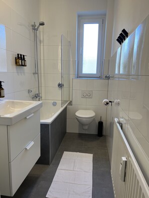 Ein modernes Badezimmer mit Badewanne, Toilette und Waschbecken. Ein Fenster sorgt für natürliches Licht, und am Boden liegt ein Handtuch.