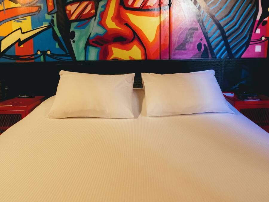 Een hotelkamer met een bed, witte kussens en een kleurrijke muurschildering.
