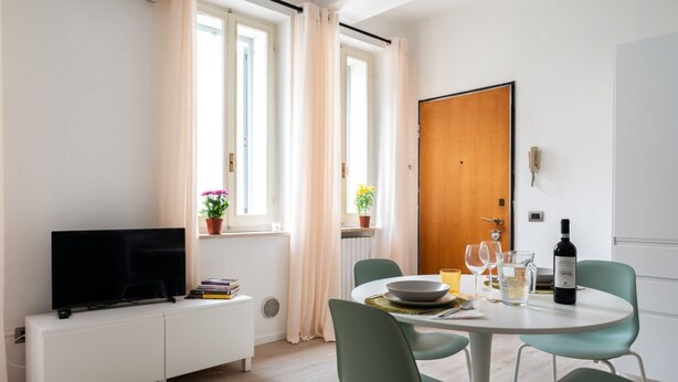A modern living room with a flat-screen TV, a dining table with chairs, a wooden door, and a window with curtains.