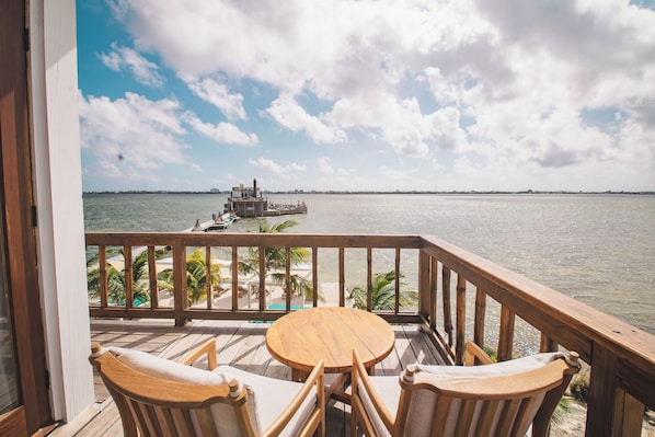 A wooden deck with a round table and two chairs, overlooking a calm body of water and a distant shoreline with palm trees.