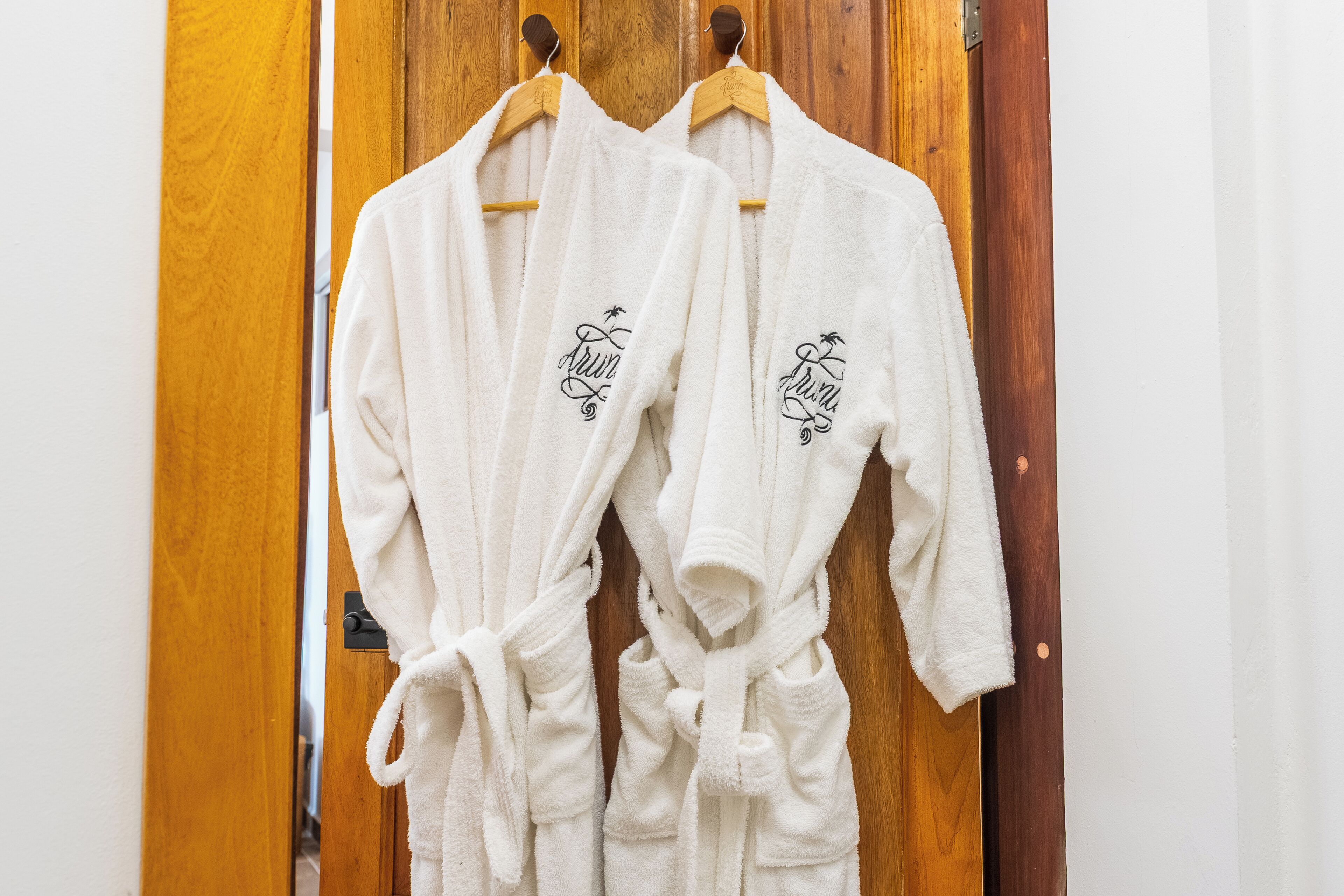 Dois roupões brancos pendurados em cabides de madeira, em frente a uma porta de madeira.