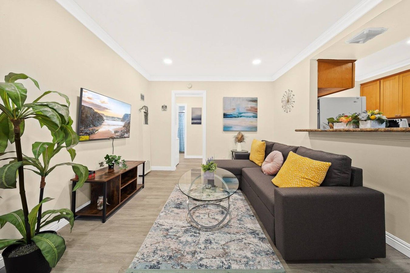 A modern living room with a dark grey sofa, a glass coffee table, and a large flat-screen TV. There are two potted plants and a wooden entertainment unit.