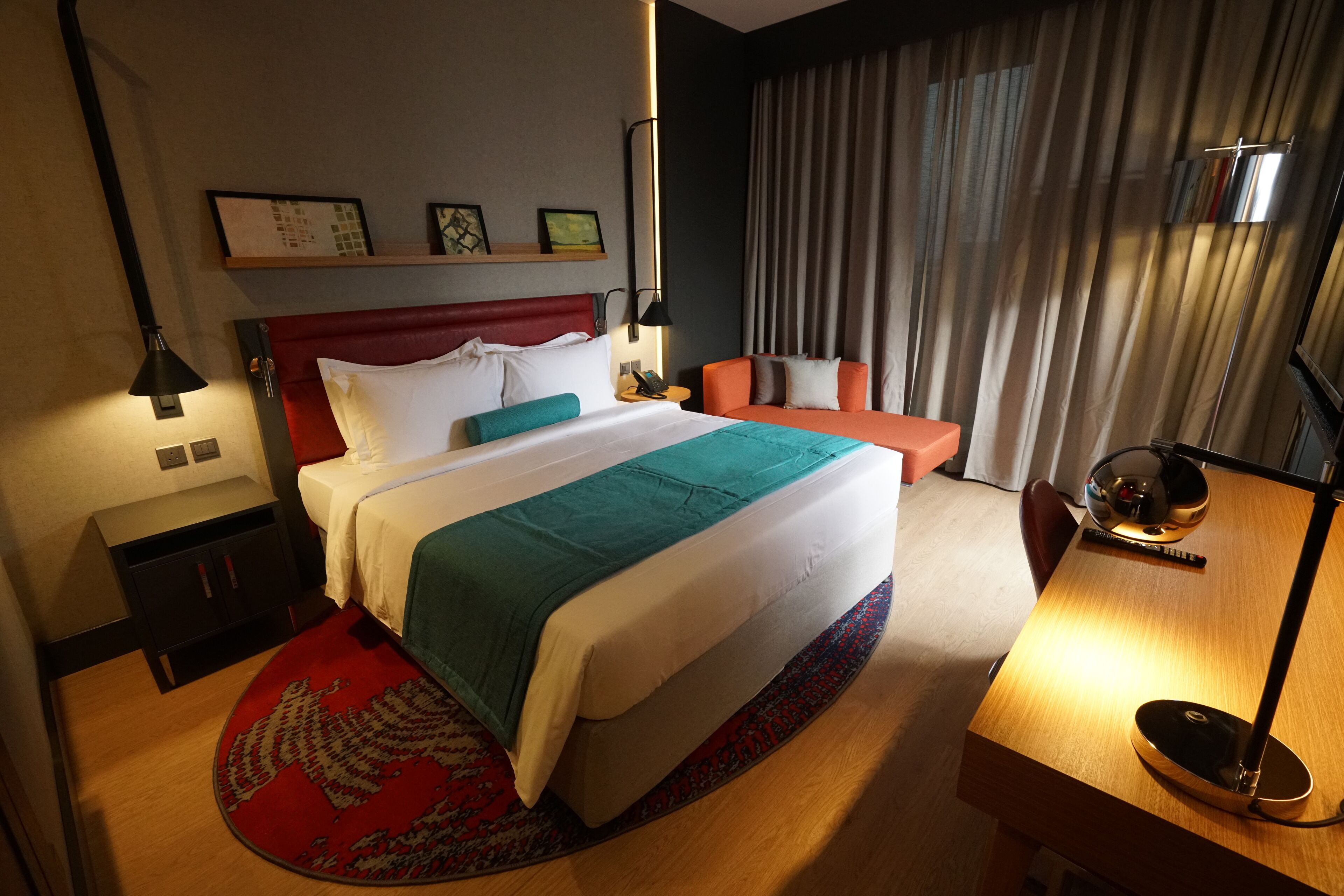 A hotel room with a large bed, a desk, a chair, a desk lamp, a nightstand, a red and white headboard, a red and white throw blanket, a red and white pillow, a red and white blanket, a red and white rug, a wooden floor, a wall-mounted shelf with pictures, a window with curtains, and a wall-mounted lamp.