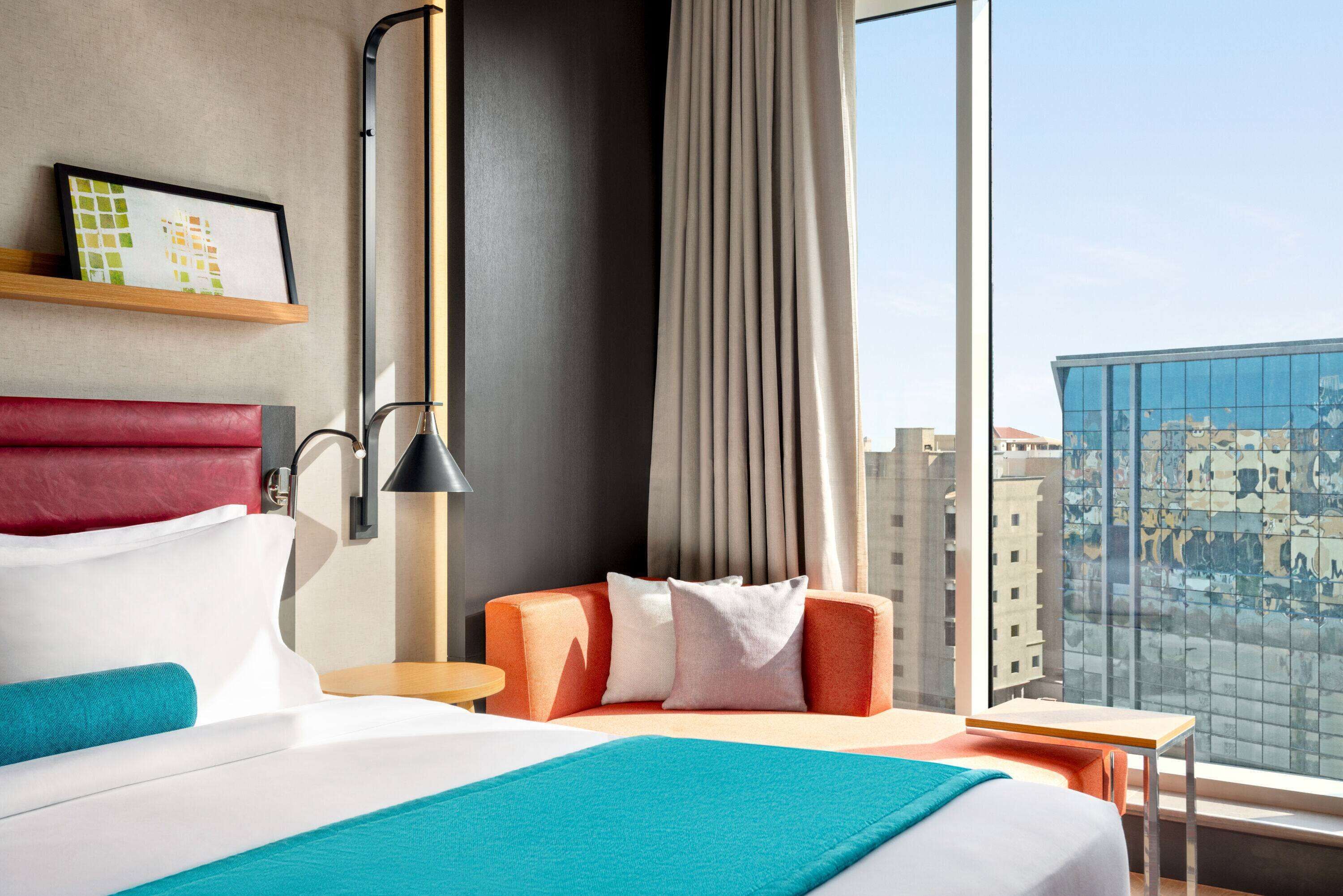 A hotel room with a large bed, a desk, a chair, a nightstand, a lamp, a window with a city view, and a framed picture on the wall.