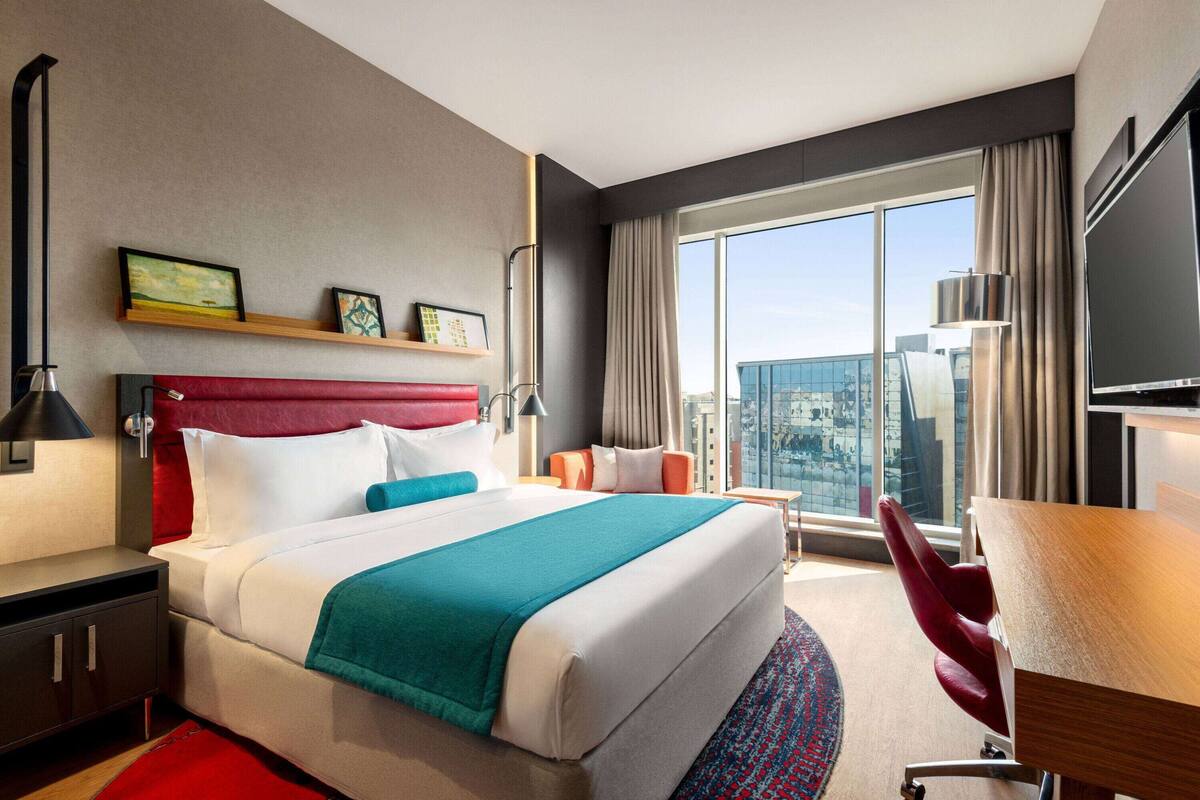 A hotel room with a large bed, a desk, a chair, a TV, and a window with a city view.