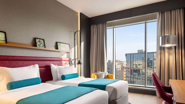 Habitación de hotel con dos camas, cabecero rojo, una ventana grande con vista a la ciudad, un escritorio y una silla.