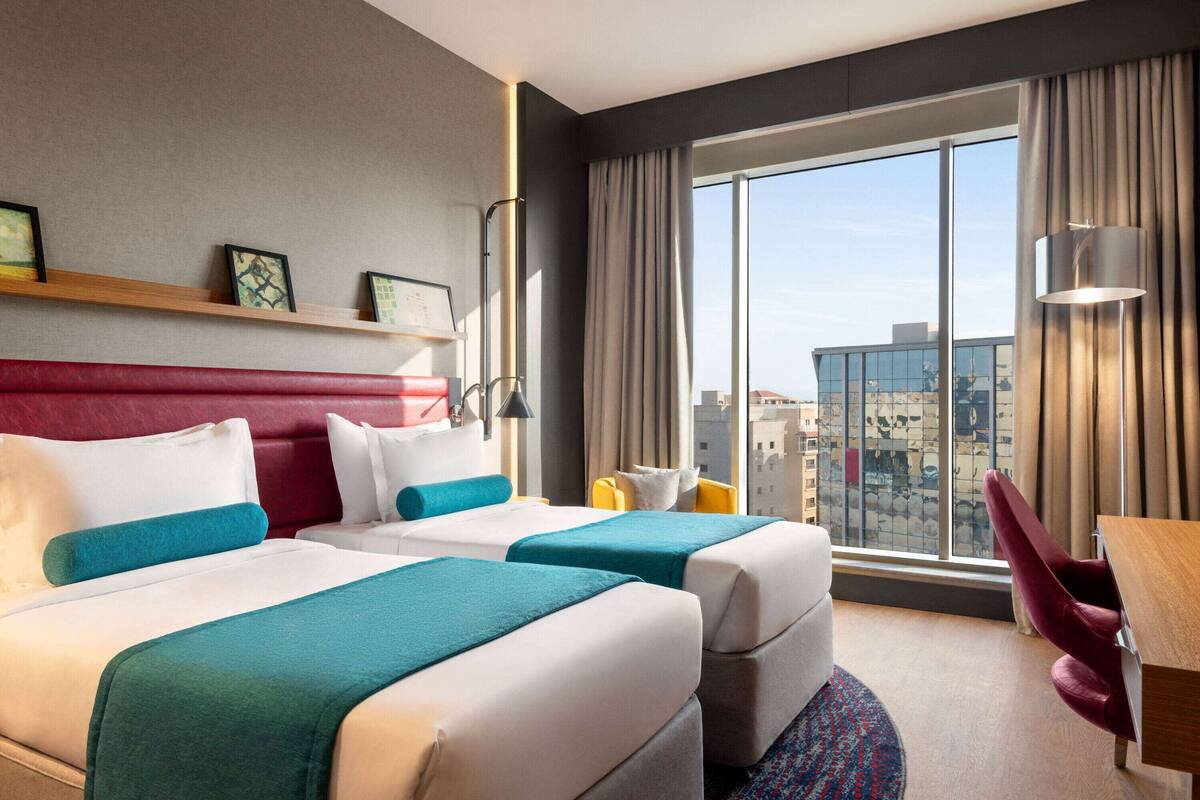 A hotel room with two beds, a red headboard, a large window with a city view, a desk, and a chair.