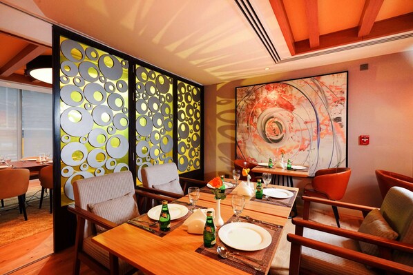 A dining area with a wooden table set for four, surrounded by chairs. A decorative panel with circular patterns is behind the table. - Ramada Encore Wyndham AL Khobar Corniche (Al Khobar)