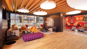 Un lobby de hotel moderno con vigas de madera en el techo, ventanas amplias y un mural llamativo.