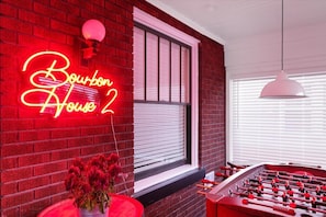 A room with a neon sign reading "Bourbon House 2", a foosball table, and a window with blinds.