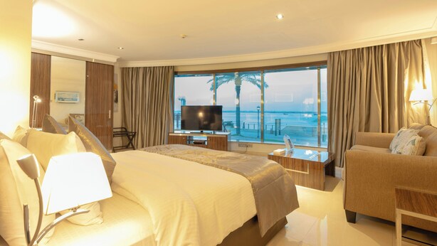 Ein Hotelzimmer mit Bett, Fernseher, Sofa, Schreibtisch und einem großen Fenster mit Meerblick.