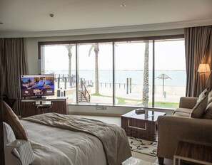 A hotel room with a bed, a TV, a desk, a chair, and a large window with a view of the sea and palm trees. - TIO SEA Resort AL Khor (Al Khor)
