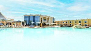 Ein großer Swimmingpool mit Liegestühlen und Sonnenschirmen, ein Gebäude mit Bögen und einer Glasfassade sowie ein klarer, blauer Himmel.
