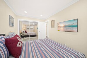 Une chambre à coucher avec un lit, une porte donnant sur une autre pièce, un téléviseur fixé au mur et un tableau représentant une scène de plage.
