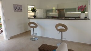 Een moderne keuken met een centraal eiland, twee barkrukken, een rieten poef en uitzicht op de aangrenzende woonkamer.