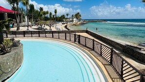 Zwembad van een resort met een stenen muur en houten omheining, met uitzicht op een strand met palmbomen en een helderblauwe lucht.