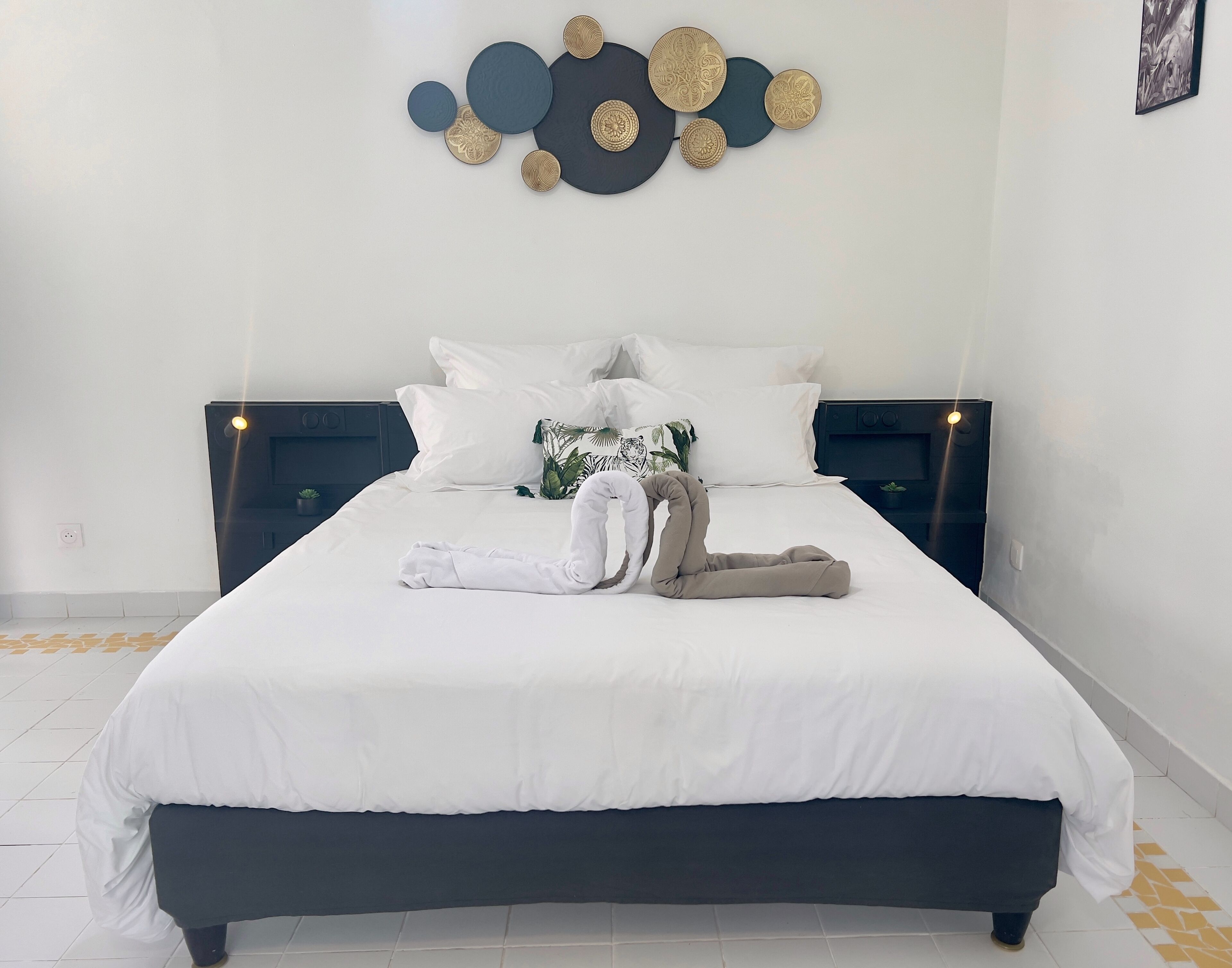 Un lit bien fait, avec une literie blanche et un coussin décoratif, une œuvre d’art accrochée au mur au-dessus du lit, et une petite plante posée sur la table de chevet.