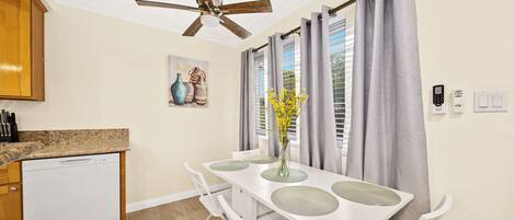 A compact kitchen with a dishwasher, a ceiling fan, a dining table with chairs, and a window with curtains.