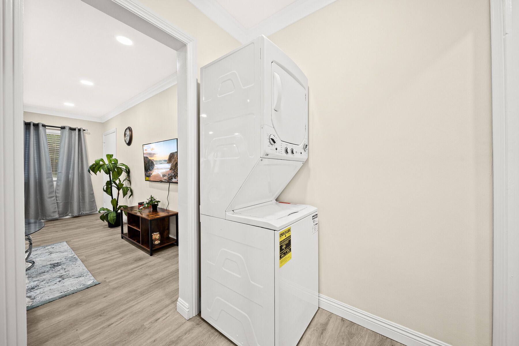 A compact laundry room with a front-loading washer and dryer, a small wooden table with a plant, and a TV mounted on the wall.