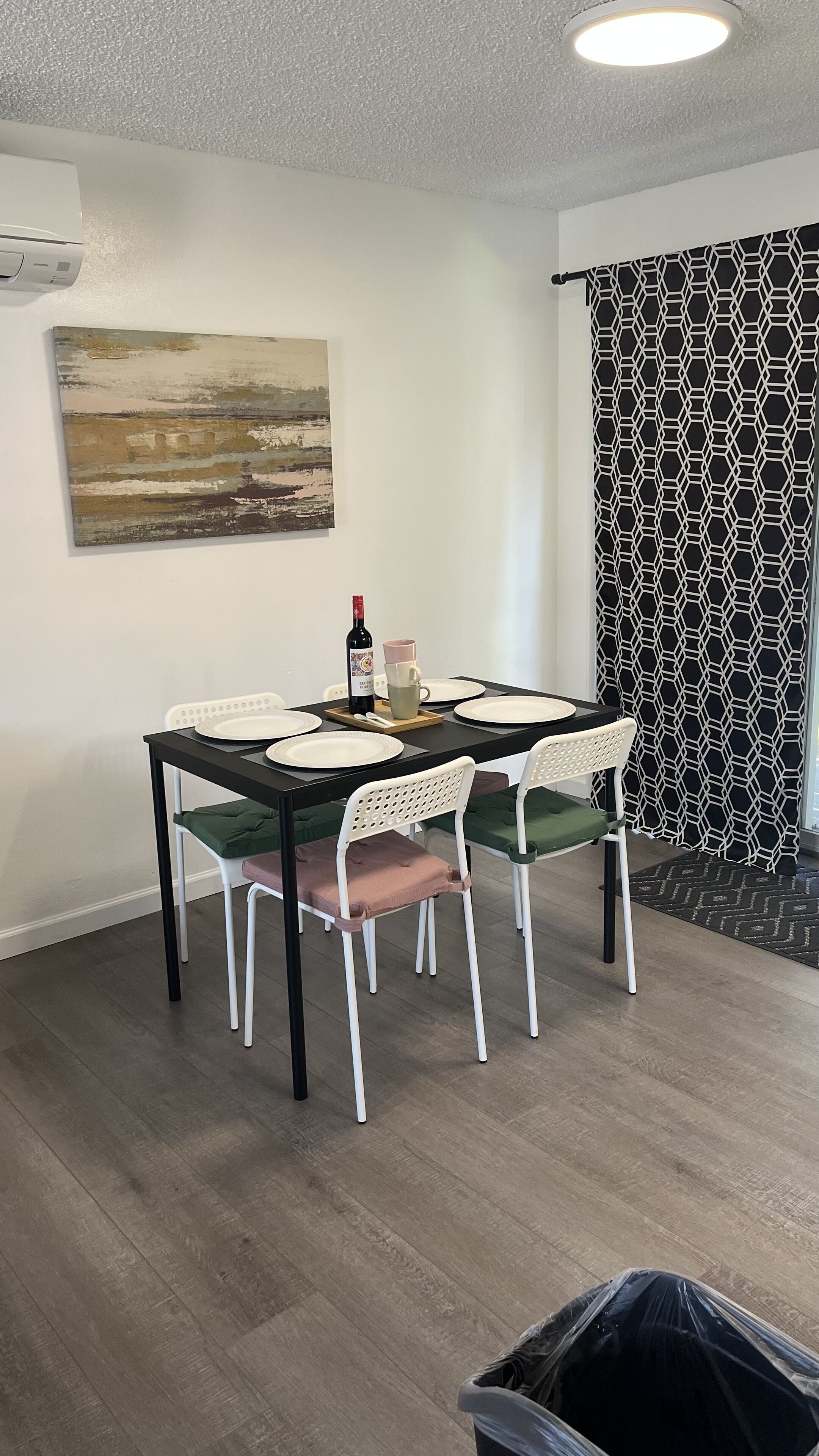 A dining area with a table set for four, white chairs with green cushions, a painting on the wall, and a patterned curtain.