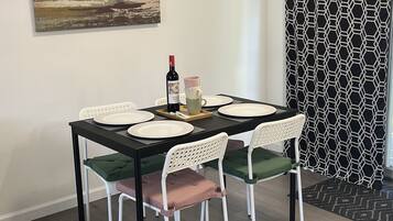 A dining area with a table set for four, white chairs with green cushions, a painting on the wall, and a patterned curtain.