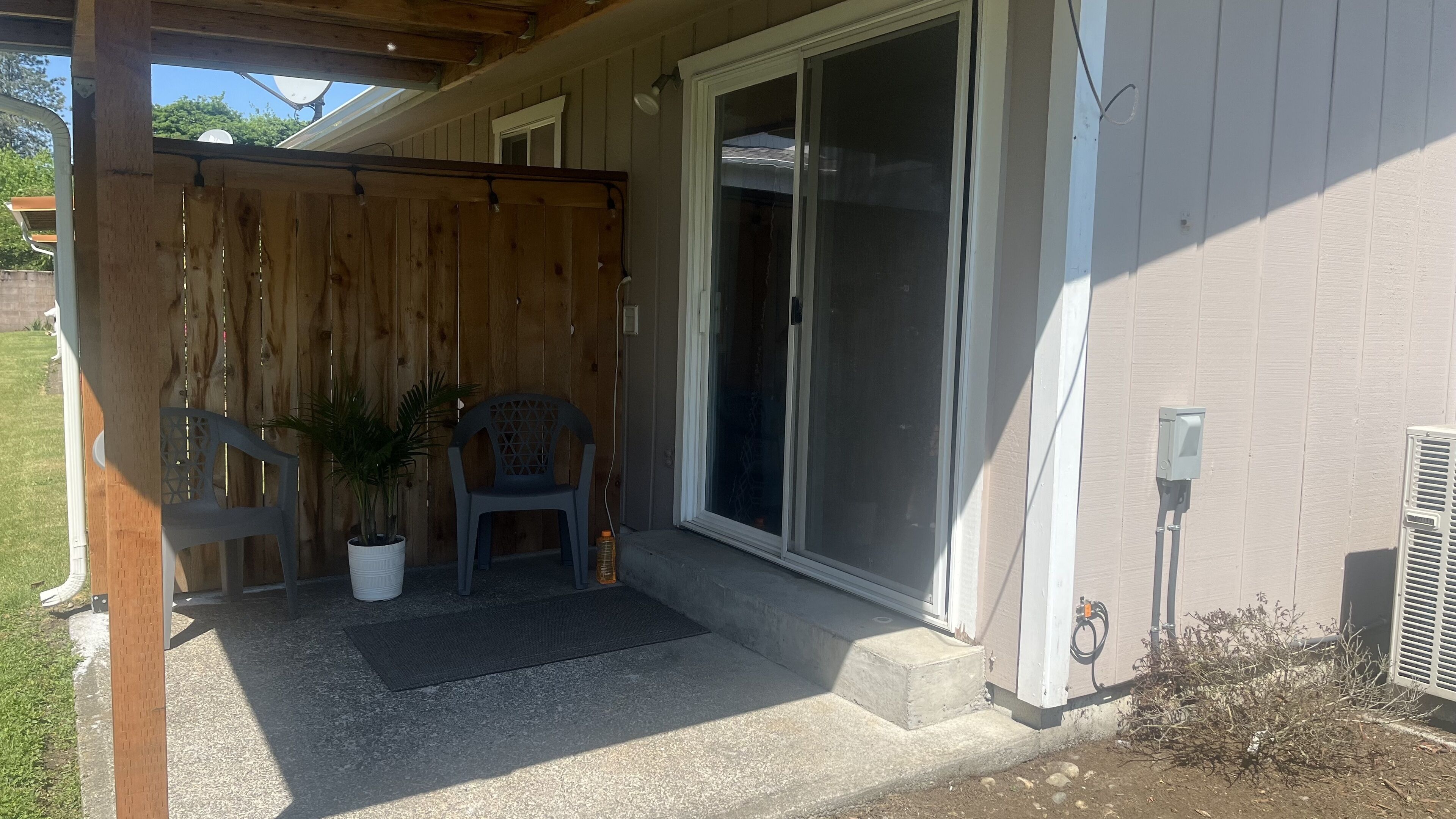 A covered porch with a wooden fence, a potted plant, a plastic chair, and a sliding glass door leading to a house.