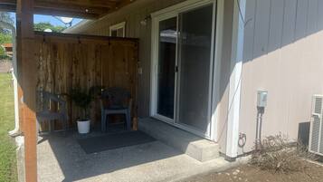 A covered porch with a wooden fence, a potted plant, a plastic chair, and a sliding glass door leading to a house.