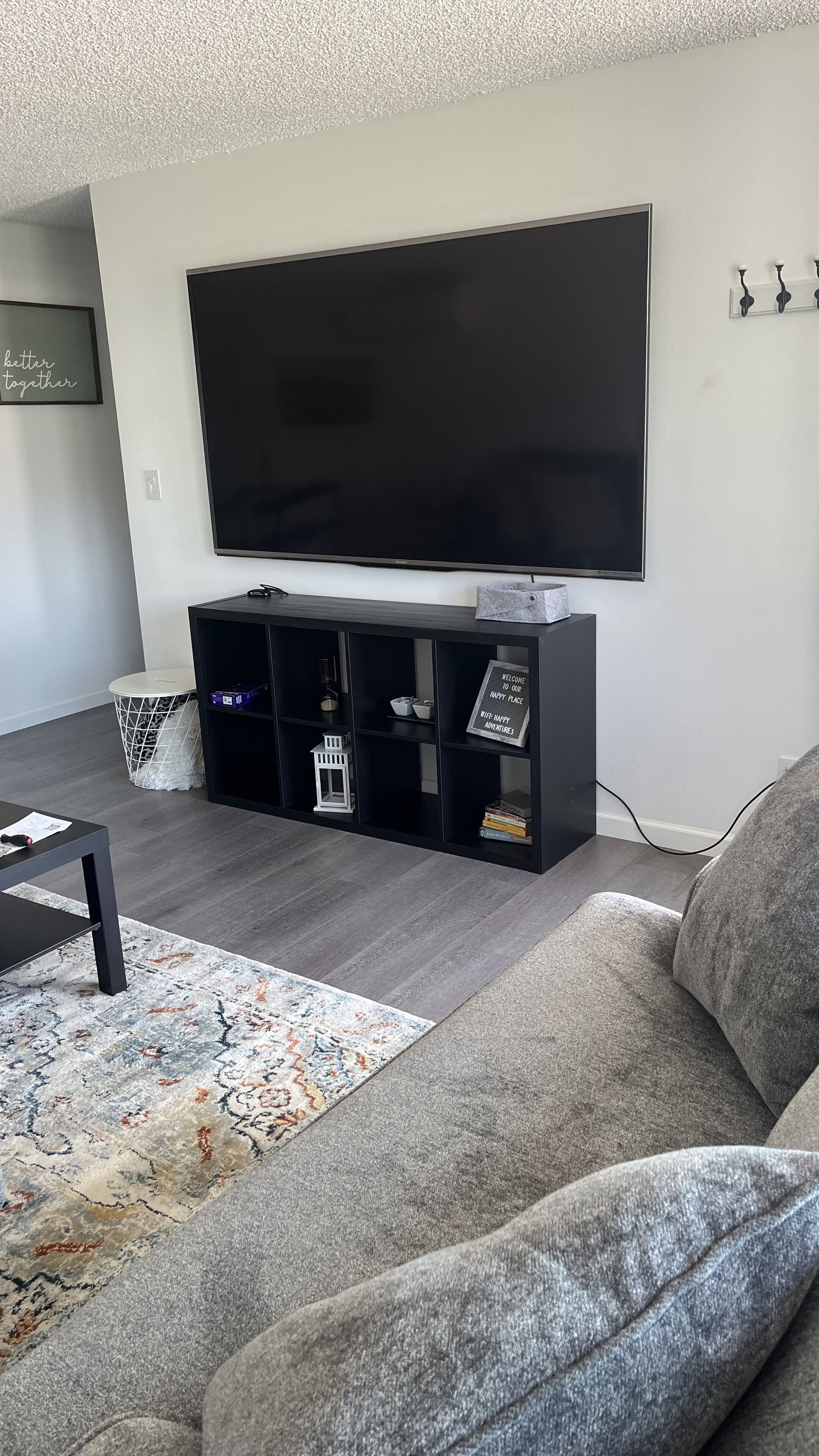 A living room with a large flat-screen TV mounted on the wall, a black entertainment unit below it, a grey sofa, and a patterned rug on the floor.