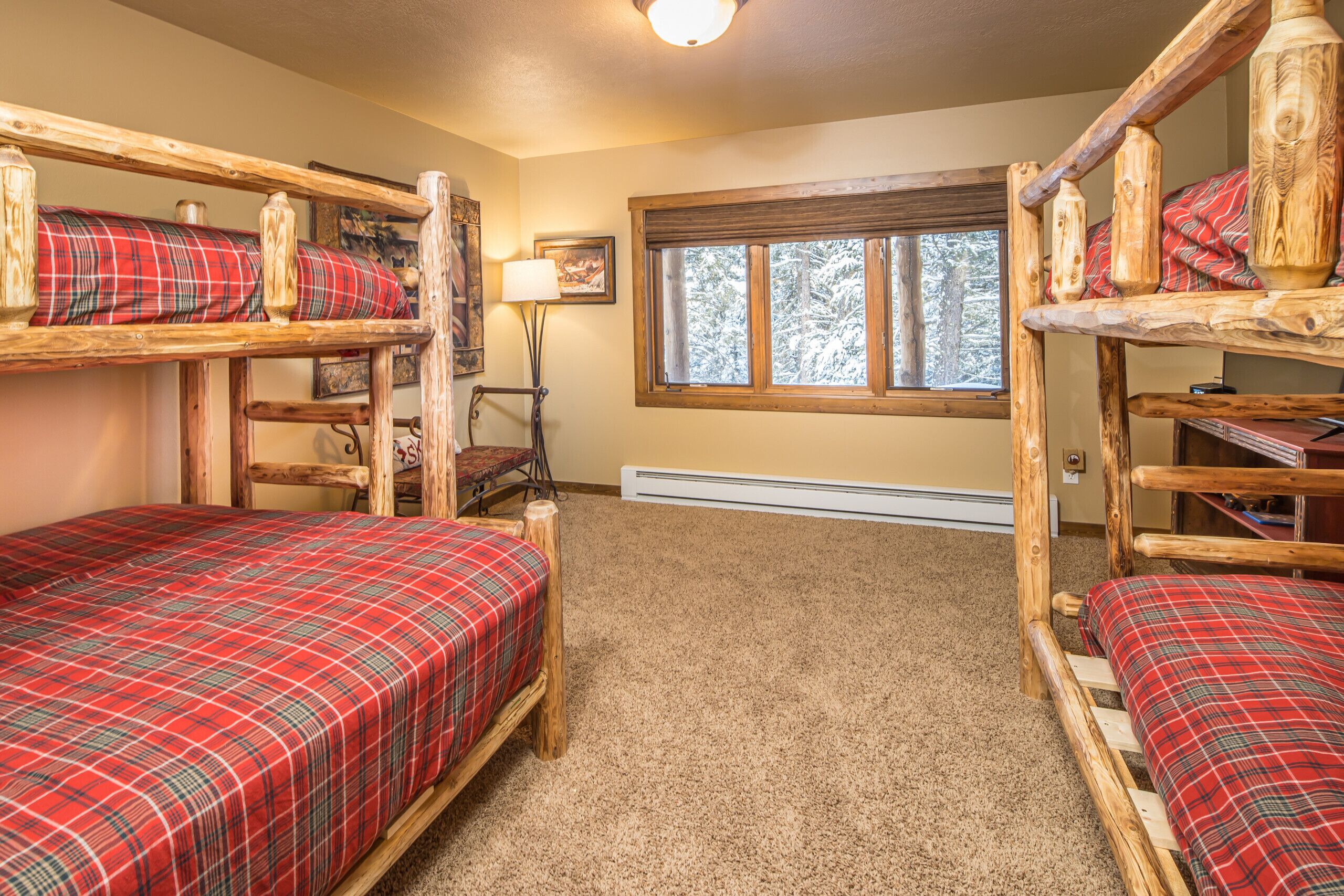 A room with three bunk beds, each with a plaid blanket. There is a window with a view of trees, a lamp, and a heater.