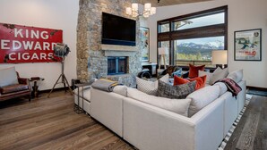 A modern living room with a stone fireplace, a large flat-screen TV, and a view of mountains through a large window. - Luxury Big Sky Ski Retreat - Moonlight Basin (Big Sky)