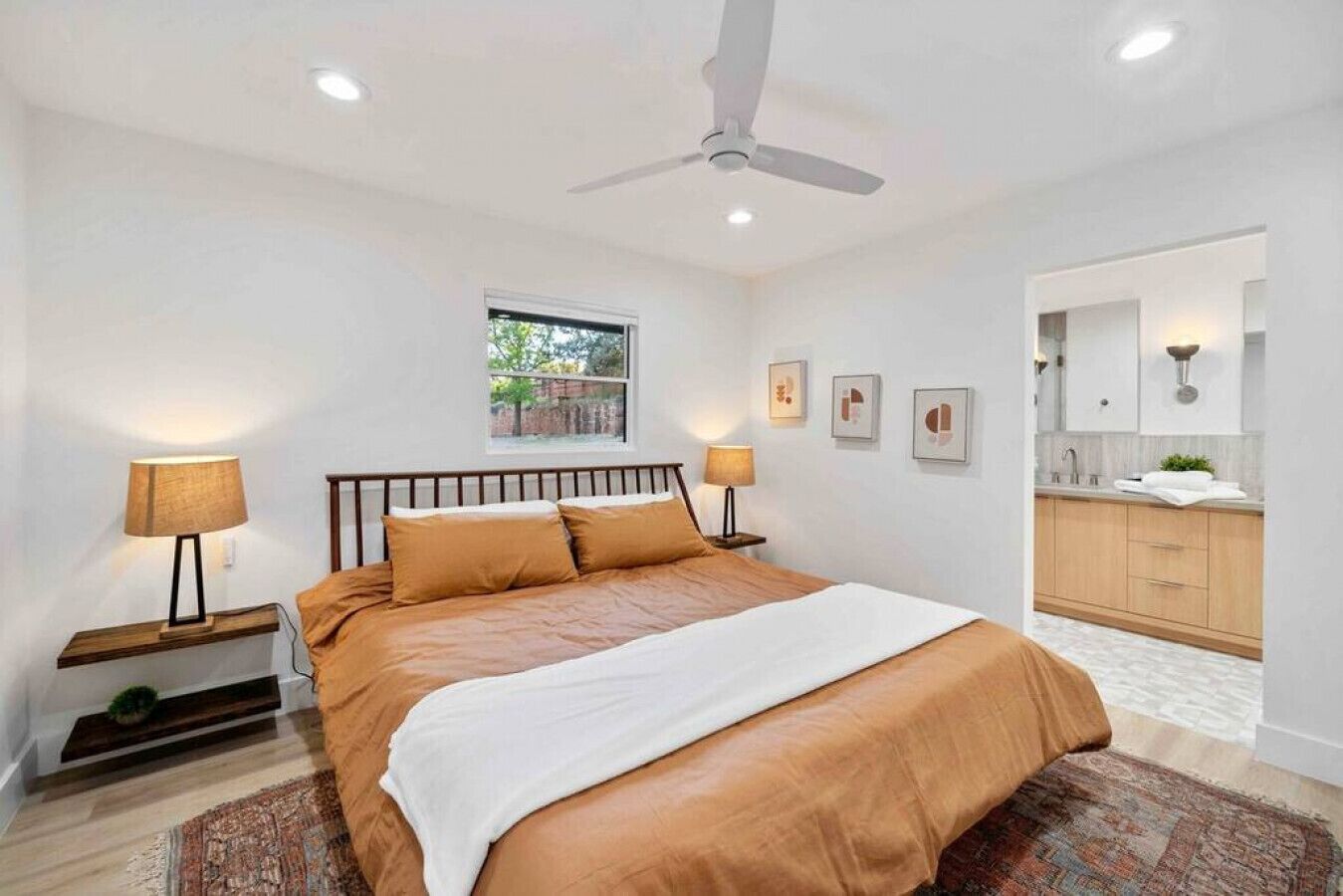 A bedroom with a bed, bedside lamps, a ceiling fan, a window, and a view of a bathroom.