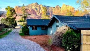 Une maison avec une allée en gravier menant à un bâtiment bleu, entourée d’arbres et avec des montagnes en arrière-plan.