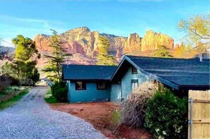 Une maison avec une allée en gravier menant à un bâtiment bleu, entourée d’arbres et avec des montagnes en arrière-plan.