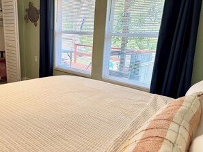 A bedroom with a bed, a window with blinds, a wall decoration, and a view of the outside.