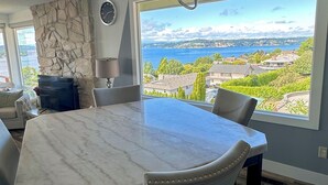Een moderne eetruimte met een grote marmeren tafel, lederen stoelen, een geaccentueerde stenen wand en uitzicht op een meer en gebouwen.