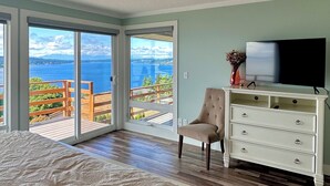 Een slaapkamer met een groot bed, een televisie, een dressoir en een stoel. Er is uitzicht op de oceaan door grote schuifdeuren.
