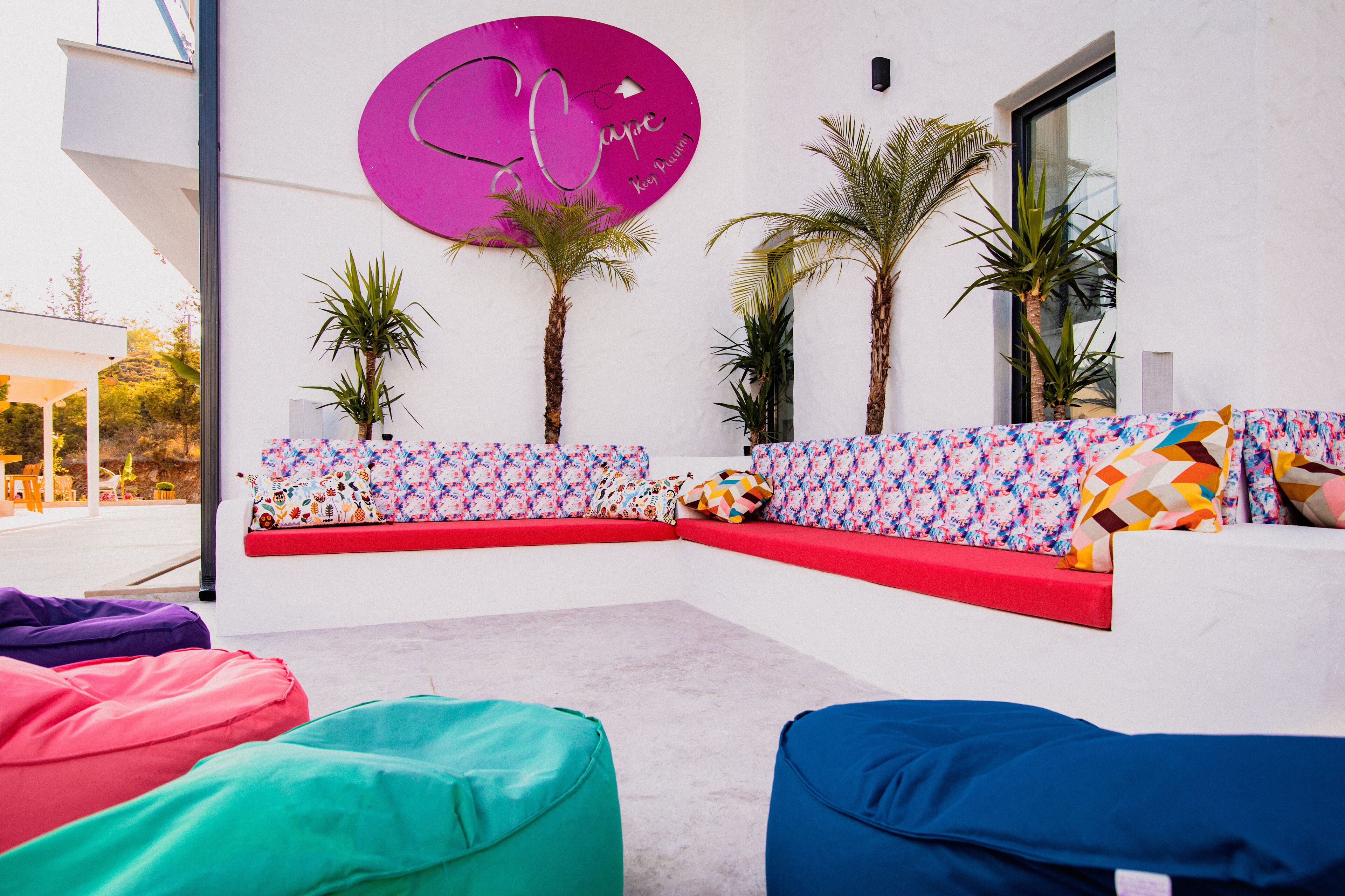 A modern outdoor seating area with colorful cushions, a vibrant pink sign, and palm trees.