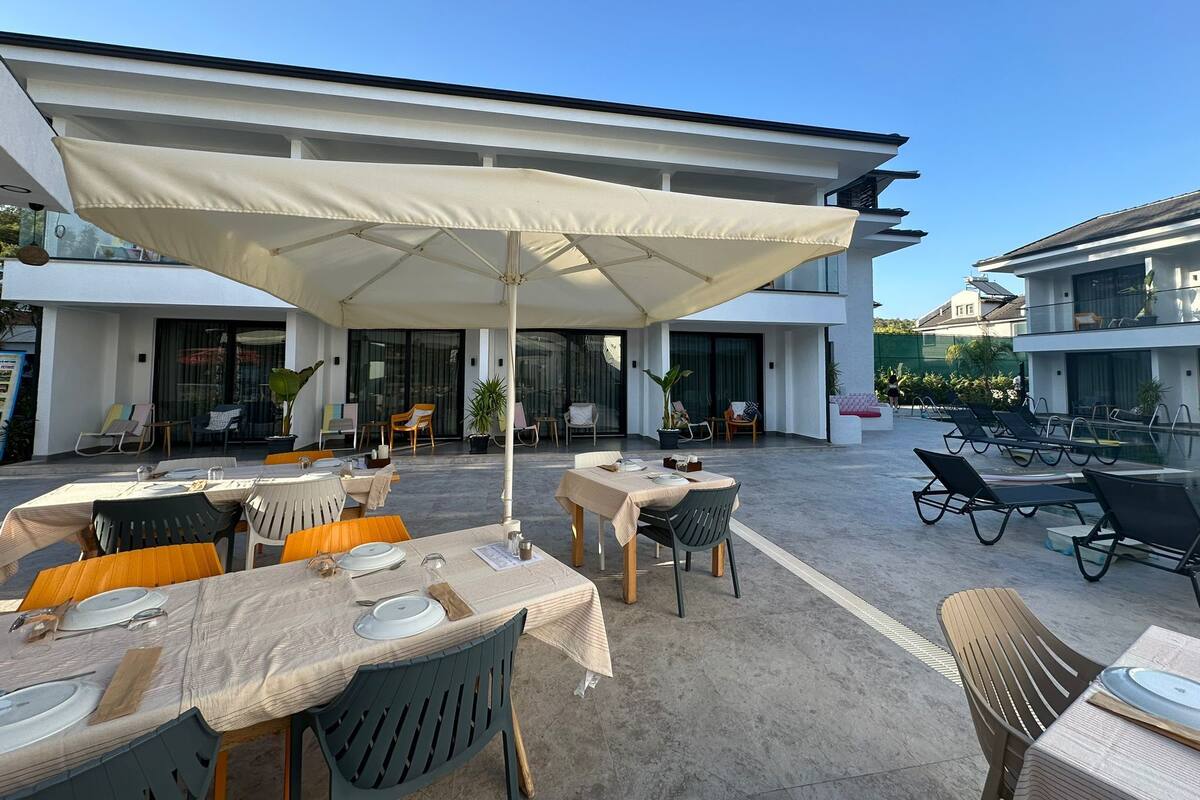 Un espace de restauration en plein air, avec des tables et des chaises, abrité par un grand parasol et entouré d’immeubles modernes.