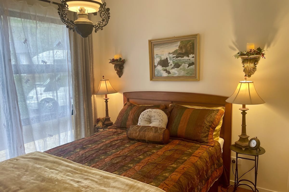 Una cama bien hecha con una colcha estampada, dos lámparas de noche, un cuadro en la pared y una ventana con cortinas translúcidas.