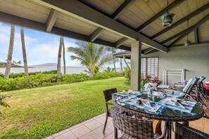 A covered outdoor seating area with a table and chairs, overlooking a grassy landscape and a body of water. - Napili Lani 318 ~ Gorgeous Napili Bay 1bd/1ba (Lahaina)