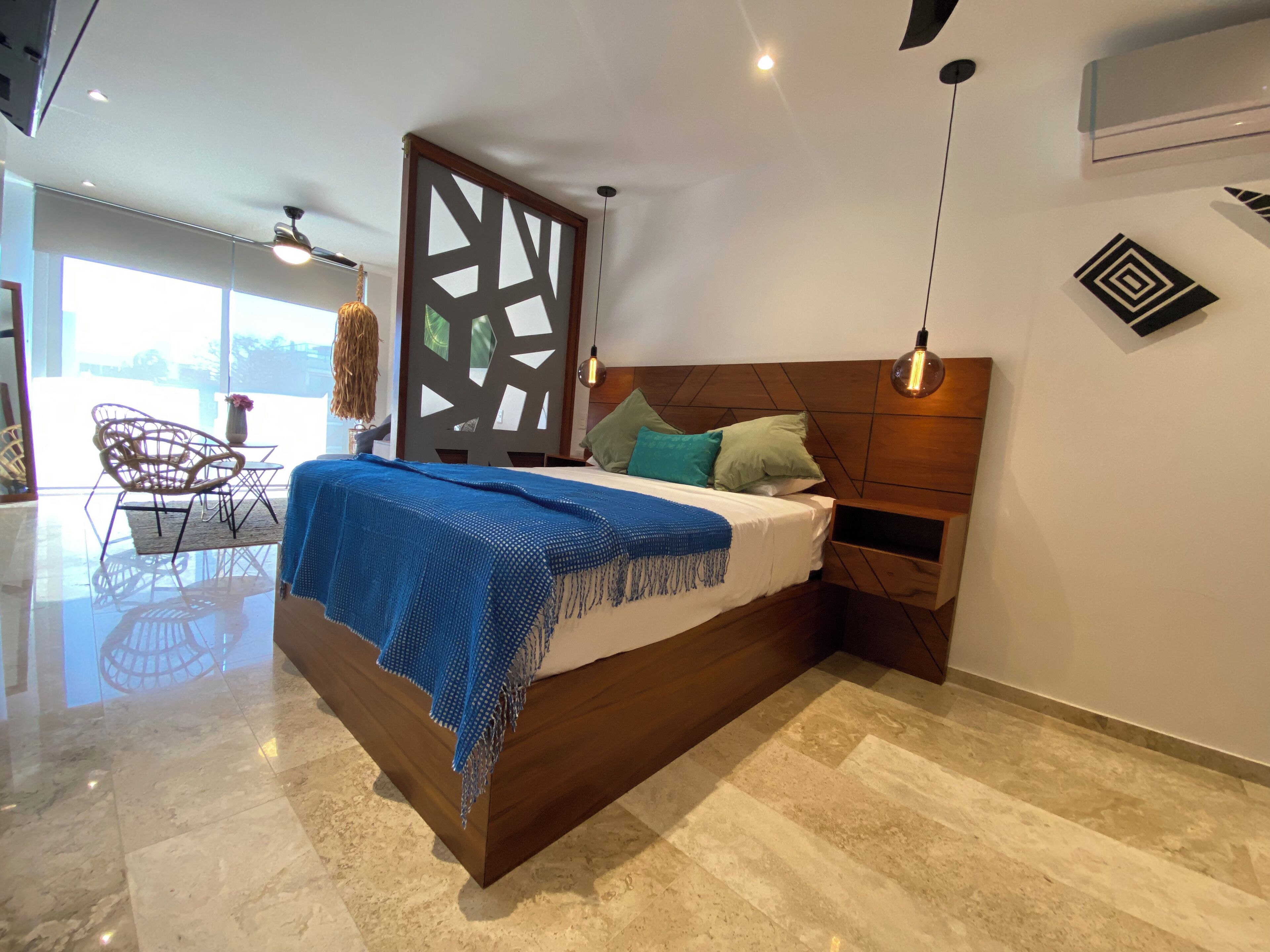 A modern bedroom with a wooden bed, a decorative partition, a hanging light fixture, and a view of the outdoors.